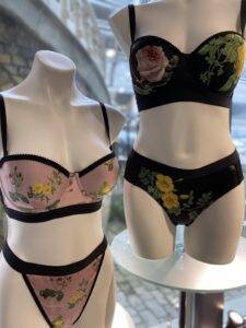 Two mannequins displaying floral lingerie sets. The left mannequin wears a light pink bra and matching underwear with a yellow flower pattern and black trim. The right mannequin has a black bra and matching underwear with a similar floral pattern featuring larger yellow and pink flowers. The designs have a classic, decorative look with floral motifs.