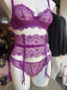 A mannequin dressed in a purple lingerie set with a sheer lace bra, a high-waisted garter belt, and matching panties. The set has a polka-dot mesh pattern and lace detailing along the edges, with thin straps and small bows on the bra and garter belt.