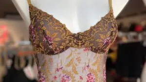 A mannequin wearing a sheer bodysuit in a warm brown color with intricate floral embroidery in gold and pink. The bodysuit has delicate lace detailing along the cups and torso, with thin shoulder straps. The embroidered floral patterns add an elegant, decorative touch to the design.