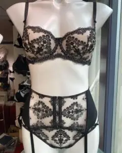 A black lace lingerie set displayed on a mannequin, featuring an underwire bra with detailed black floral lace and a matching high-waisted garter belt with intricate lace patterns. The garter belt includes straps for attaching stockings, creating a vintage-inspired, elegant look.