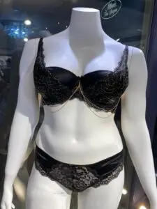 A black bra and panty lingerie set displayed on a mannequin. The bra features lace detailing with delicate gold chain accents draping along the cups, while the matching panties have floral lace across the front, adding elegance to the satin fabric.