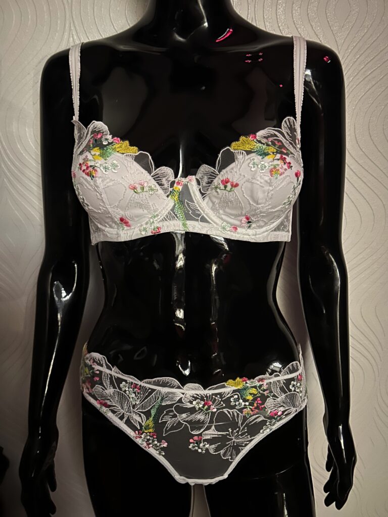 A glossy black mannequin wearing a white lace lingerie set with colorful floral embroidery. The bra has sheer lace cups adorned with pink, yellow, and green floral details, while the matching panties feature similar embroidery with a scalloped lace trim. The mannequin is positioned against a textured white wall background.