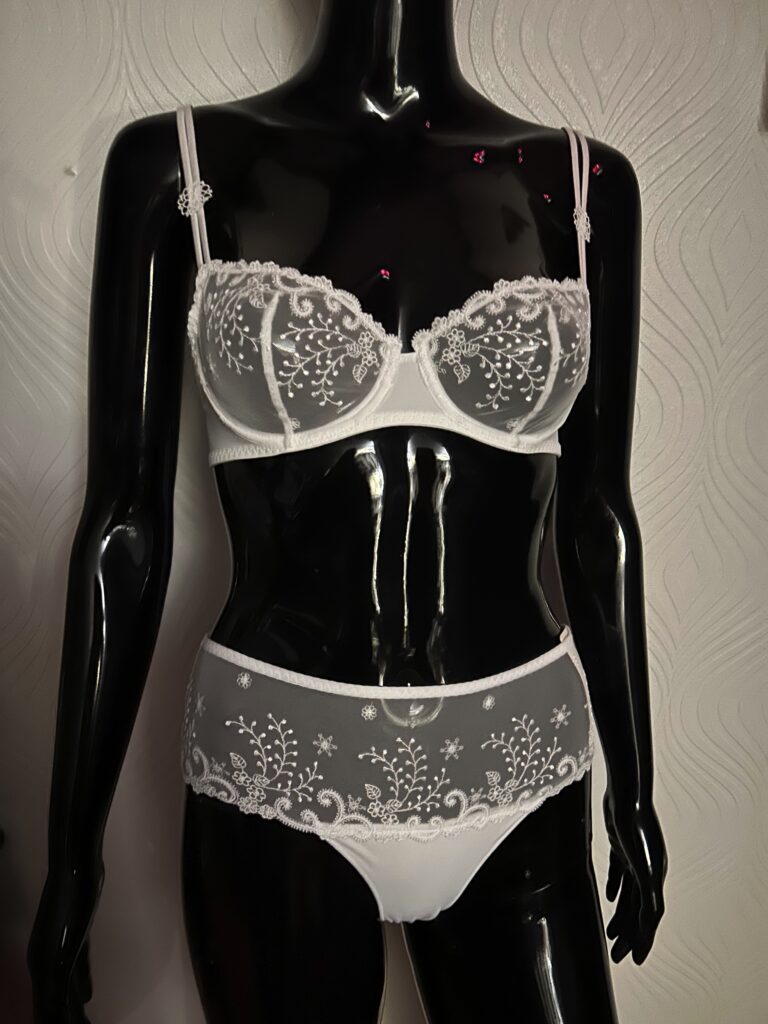 A glossy black mannequin wearing a white lace lingerie set. The bra features intricate sheer lace with floral embroidery and a small satin bow in the center. The matching panties have a combination of lace and opaque fabric with delicate embroidery along the waistband and sides. The mannequin is positioned against a textured white wall background.