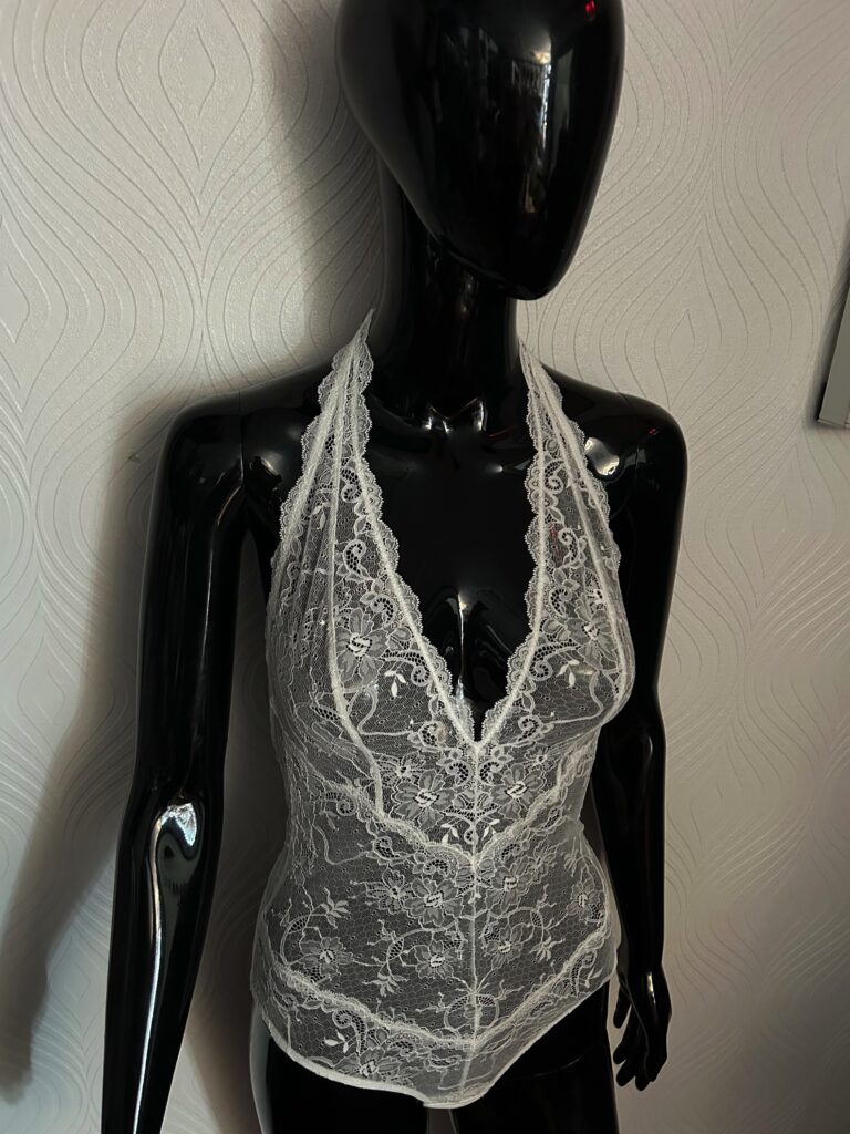 A glossy black mannequin wearing a white lace bodysuit with a halter neckline. The sheer lace fabric features an intricate floral pattern, with scalloped lace trim along the neckline and edges. The mannequin is posed against a textured white wall background.